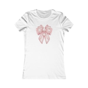 Puede incluir: Camiseta blanca con un diseño de lazo rosa con detalles de perlas. El lazo tiene una apariencia delicada y de encaje y está centrado en la parte delantera. La camiseta tiene cuello redondo y mangas cortas.