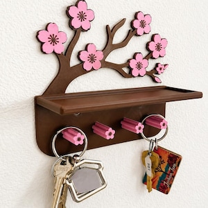 以下が含まれることがあります： 茶色の木の枝のシルエットとピンクの桜の花が特徴の壁掛けキーホルダー。小さな棚と鍵を掛けるための4つのピンクの花の形をしたフックが付いており、フックには2つのキーリングが取り付けられています。