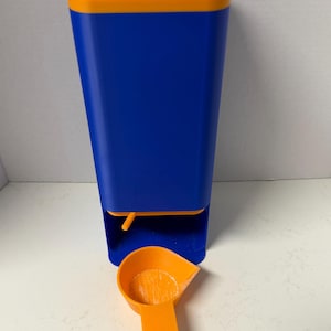 May include: A blue and orange pet food dispenser with a matching scoop. The dispenser is rectangular with a square top and a small opening. The scoop is orange and has a handle.
