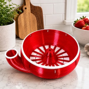 Puede incluir: Un descorazonador de fresas circular de color rojo brillante con un cono central, un borde blanco y un pico lateral. El dispositivo tiene una base interior con ranuras diseñada para procesar fresas, con un acabado brillante y funcional.