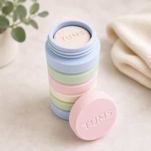 May include: A cylindrical stack of pastel-colored, circular Tums antacid tablets in shades of light blue, pale green, and soft pink. The tablets are arranged vertically, with one pink tablet resting upright in front of the stack, clearly displaying the embossed text TUMS on its surface.