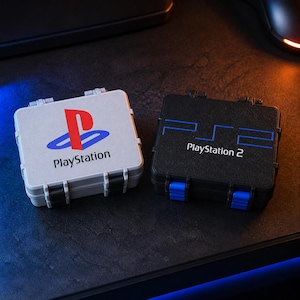 May include: Two square, hinged storage cases. The left case is light gray with the red, blue, and yellow PlayStation logo and text. The right case is black with the blue PS2 logo and the text PlayStation 2, featuring bright blue latch accents.