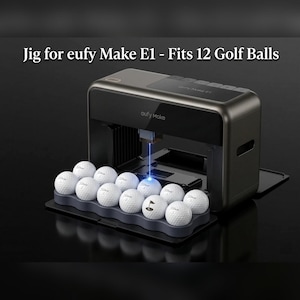 May include: A dark gray Eufy Make E1 machine with a tray holding 12 white golf balls. The text "Jig for eufy Make E1 - Fits 12 Golf Balls" is displayed above. A blue laser beam points from the machine onto one of the golf balls in the tray.