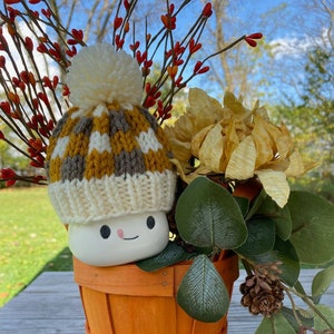 May include: A white ceramic marshmallow with a brown and white knit hat and a pom pom sits in an orange wooden bucket. The bucket is surrounded by fall foliage, including yellow flowers, green leaves, and red berries.