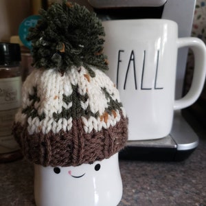 May include: A white ceramic mug with a smiling face, wearing a brown, green, and white knitted hat with a green pom-pom. The mug is sitting on a brown countertop next to a white mug with the word "FALL" written on it.