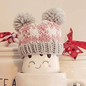 May include: A pink and gray knitted mug cozy with two gray pom-poms on top. The cozy is on a white mug with a smiley face.