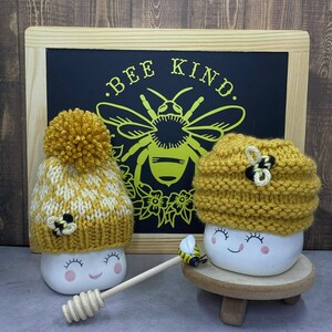 May include: Two white ceramic mugs with smiling faces, each wearing a knitted yellow and white bee-themed hat. The mugs are sitting on a wooden surface in front of a chalkboard with the text "Bee Kind" and a bee illustration.