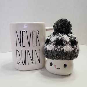 May include: White ceramic mug with black text that reads "NEVER DUNN". A white ceramic marshmallow with a black and white knit hat sits next to the mug.