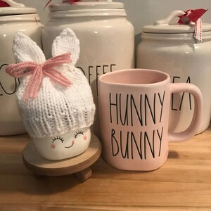 May include: A white ceramic mug with the text "Hunny Bunny" in black lettering. The mug is sitting on a wooden surface next to a white ceramic jar with a red and white polka dot ribbon. A white knitted bunny hat with pink ears and a pink ribbon is sitting on a small wooden stand.