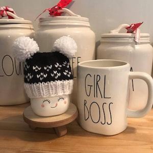 May include: White ceramic mug with black text that reads "Girl Boss". A white ceramic jar with a red ribbon is in the background. A white marshmallow with a black and white knitted hat with white pom-poms is on a wooden stand.