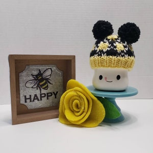 May include: A white ceramic mug with a happy face and black eyes, wearing a yellow and black knit hat with black pom-poms. The mug is on a blue cake stand with a yellow felt flower.