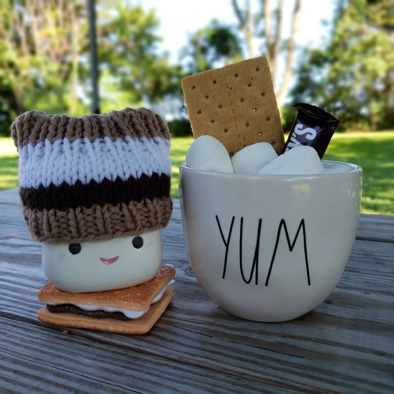May include: A white ceramic mug with the word "YUM" printed on it, filled with marshmallows and graham crackers. A knitted brown and white hat sits on top of a white marshmallow with a chocolate bar and graham crackers.