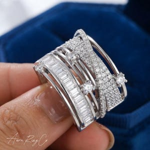 May include: A wide silver-toned ring featuring a multi-band design. One section displays a row of rectangular baguette-cut stones, while another section is covered in small round pavé-set stones. Two larger round stones are set between the bands.