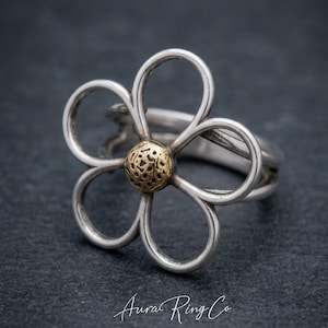 May include: A silver flower ring with a gold-coloured centre. The ring features a floral design with five curved petals. The ring is set against a dark background. The text "Aura Ring Co" is at the bottom.