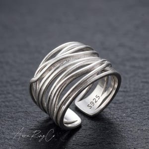 Puede incluir: Un anillo plateado con un diseño tejido. El anillo presenta múltiples hebras de metal que se enrollan alrededor del dedo. El interior del anillo está estampado con "S925". El anillo se muestra sobre una superficie oscura.