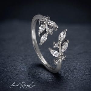 Peut inclure: Une bague ouverte argentée composée de deux branches en forme de feuilles ornées de pierres transparentes taille marquise et rondes. L'anneau porte le poinçon 925 et le texte Aura Rings Co est visible dans le coin inférieur gauche de l'image.