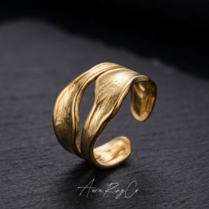 May include: A gold-colored ring with a textured, organic design. The ring features a split band with two curving, leaf-like elements that overlap. The ring is set against a dark background, with the text "Aura Ring Co" in the bottom right corner.
