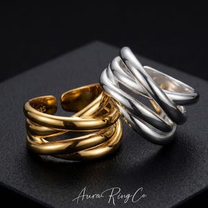 May include: Two adjustable rings, one gold and one silver, are shown on a black surface. The rings have a multi-band design, with the gold ring on the left and the silver ring on the right. The rings are from Aura Ring Co.