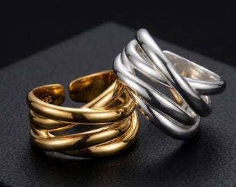 Può includere: Due anelli regolabili, uno dorato e uno argentato, sono esposti su una superficie nera. Gli anelli presentano un design a più bande, con l'anello dorato a sinistra e l'anello argentato a destra. Gli anelli sono di Aura Ring Co.
