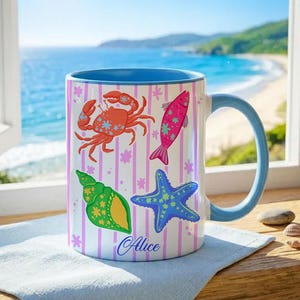 Puede incluir: Taza de cerámica blanca con interior y asa azules, decorada con criaturas marinas coloridas: un cangrejo, un pez, una concha y una estrella de mar. La taza tiene un fondo a rayas y el nombre "Alice" en cursiva.