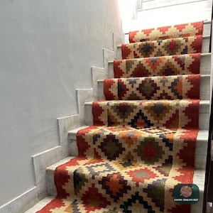 Può includere: Una lunga passatoia per scale con un motivo geometrico a rombi ripetuto nei toni del rosso, arancione, verde e grigio scuro su uno sfondo beige naturale. Il tappeto copre diversi gradini e include un piccolo logo con scritto Luxury Beneath Feet.