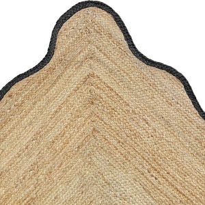 Op de afbeelding: Een hoeksectie van een geweven beige jute vloerkleed met een opvallende geschulpte rand. Het natuurlijke vezelmateriaal heeft een getextureerd, gevlochten uiterlijk en de gebogen rand is afgewerkt met een stevige zwarte stoffen bies die de golvende vorm volgt.