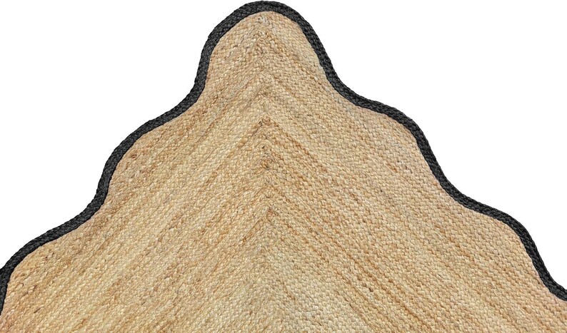 Op de afbeelding: Een hoeksectie van een geweven beige jute vloerkleed met een opvallende geschulpte rand. Het natuurlijke vezelmateriaal heeft een getextureerd, gevlochten uiterlijk en de gebogen rand is afgewerkt met een stevige zwarte stoffen bies die de golvende vorm volgt.