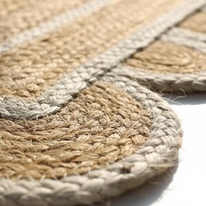 K&ouml;nnte beinhalten: Ein gewebter Teppich aus nat&uuml;rlichen, hellbraunen Jutefasern mit einem wellenf&ouml;rmigen Randdesign. Die kreisf&ouml;rmigen Muster sind mit einer dicken, geflochtenen hellgrauen Bord&uuml;re eingefasst, die sich von der strukturierten, erdbraunen Oberfl&auml;che abhebt.