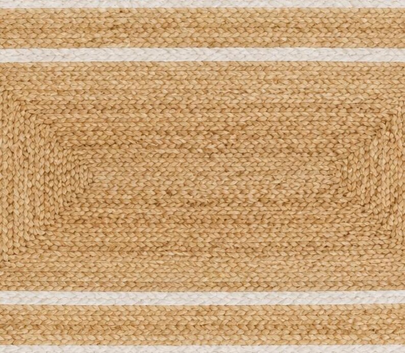 K&ouml;nnte beinhalten: Ein rechteckiger Teppich mit einer geflochtenen Textur aus nat&uuml;rlichem, hellbraunem Jute und horizontalen wei&szlig;en Streifen. Das Design besteht aus einem zentralen geflochtenen Juterechteck, umgeben von abwechselnden B&auml;ndern aus Jute und wei&szlig;em Material.