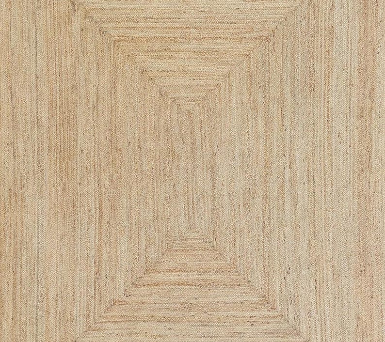 Op de afbeelding: Een rechthoekig vloerkleed gemaakt van natuurlijke bruine jutvezels, met een gevlochten constructie gerangschikt in concentrische rechthoekige patronen die vanuit het midden naar de buitenranden uitstralen, wat zorgt voor een uniform en getextureerd oppervlak.