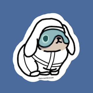 Op de afbeelding: Een gestanste sticker met een cartoonillustratie van een wit konijn dat een lichtblauw slaapmasker en een wit kledingstuk met capuchon draagt. Het konijn heeft lange oren en een beige gezicht. De tekst BIOLOGYOBSESSED is in kleine letters gedrukt.