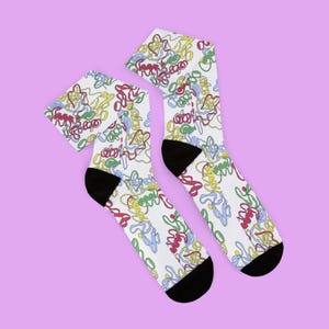 May include: A pair of white crew socks featuring a repeating pattern of abstract, interconnected squiggly lines in shades of red, yellow, green, and blue. The socks have solid black heels and toes, providing a distinct contrast to the white fabric.