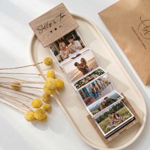 May include: A wooden photo album with a hinged lid, displaying multiple photographs. The lid is engraved with the names "Sally & Tom" and decorative symbols. The album is open, revealing a series of connected photos. The album is on a white oval tray.