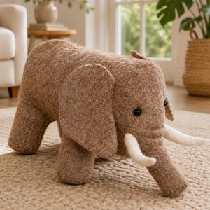 May include: A brown, textured elephant-shaped footstool with large ears, a trunk, and two white tusks. The elephant has small, dark, round eyes and a sturdy, four-legged design, crafted from a soft, fleecy material with a neutral, earthy tone.