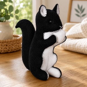 Puede incluir: Un juguete de peluche con forma de ardilla hecho de fieltro negro y blanco. La ardilla tiene una cola tupida, vientre blanco y sostiene una nuez blanca entre sus patas delanteras. Costuras blancas visibles bordean las orejas, el cuerpo y las extremidades.