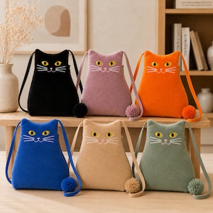 May include: Six cat-shaped crossbody bags in black, mauve, orange, blue, beige, and sage green. Each bag features embroidered yellow eyes, a pink nose, white whiskers, and a matching pom-pom tail attached to the bottom corner with a strap.