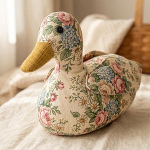 May include: A stuffed duck toy made from cream-colored fabric featuring a floral pattern of pink roses, blue hydrangeas, and white daisies. The duck has a textured mustard yellow fabric beak and a small black button eye on each side of its head.