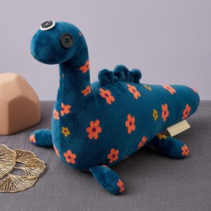Op de afbeelding: Een pluche dinosaurus van diepblauwe stof met een oranje bloemenpatroon. Het heeft twee zwarte knoopogen, een kleine witte knoop bovenop de kop en een rij zachte blauwe vinnen op de rug. Een klein beige label is zichtbaar aan de zijkant.