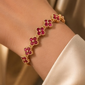 May include: A gold-toned bracelet featuring a series of connected four-leaf clover motifs. Each clover is outlined in gold metal and contains four small, vibrant red circular stones set within the petals, creating a continuous floral pattern.
