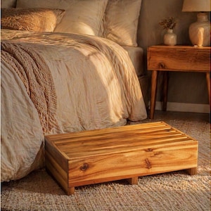 May include: A rectangular wooden step stool with a natural finish. The stool has a slatted top and is positioned in front of a bed with beige bedding. The room has a warm, inviting atmosphere, with a bedside table and lamp visible in the background.