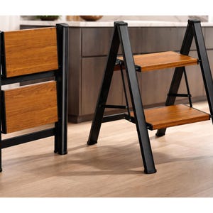 May include: A folding step stool with a black metal frame and two wooden steps. The step stool is shown both folded and unfolded, showcasing its compact design and ease of use. The wooden steps have a warm, natural wood grain finish.