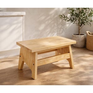 May include: A small, light-colored wooden step stool. The stool has a rectangular top and angled legs, with a supporting beam. The wood grain is visible. The stool is approximately 12 inches long, 8 inches wide, and 8 inches tall.