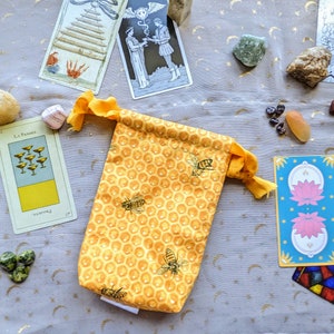 May include: A yellow drawstring pouch with a honeycomb pattern and bee illustrations. The pouch is surrounded by tarot cards, crystals, and other objects.