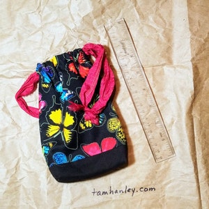 May include: A black drawstring bag with a butterfly print. The bag is made of fabric and has a red drawstring. The bag is sitting on a brown paper surface. The text "tamhanley.com" is written on the surface.