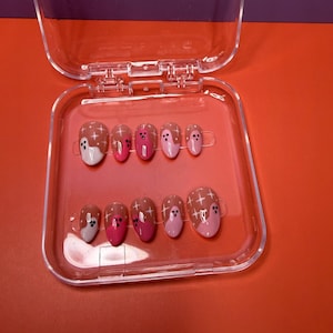 May include: A set of ten almond-shaped press-on nails displayed in a clear plastic case. The nails feature a color palette of pink, light pink, and brown, decorated with white ghost figures and small white star patterns across the surface.