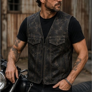 Male rider wearing a distressed dark camo motorcycle vest with a V-neck, front zipper and flap chest pockets beside a motorcycle.