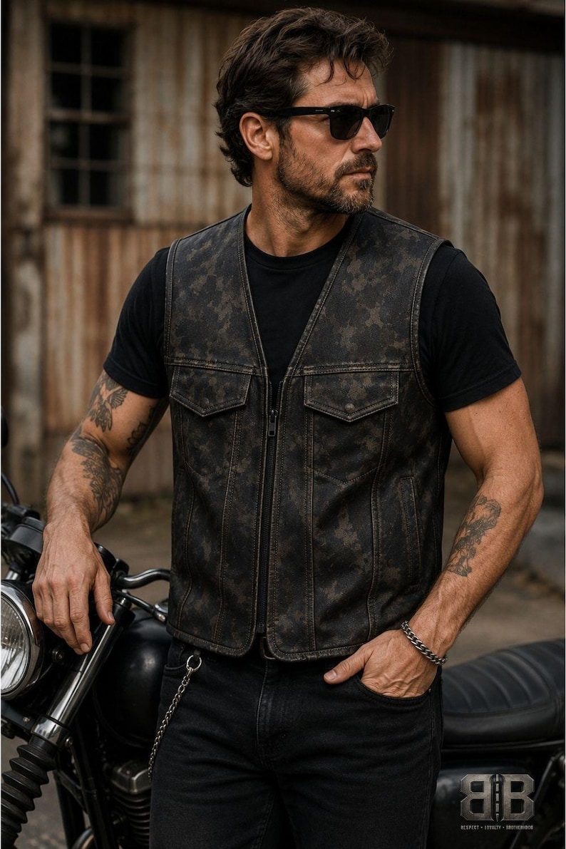 Male rider wearing a distressed dark camo motorcycle vest with a V-neck, front zipper and flap chest pockets beside a motorcycle.