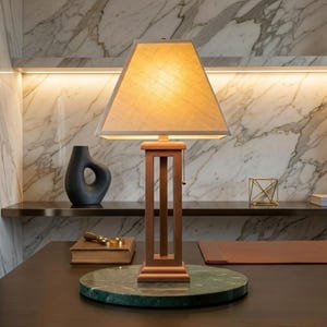 May include: A wooden table lamp with a rectangular lampshade. The lamp has a brown wooden frame and a green marble base. A black vase and a gold geometric sculpture are also on the table. The background features a marble wall.