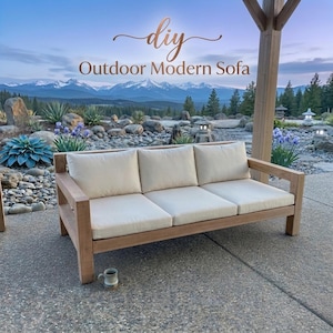 Może przedstawiać: Drewniana sofa ogrodowa z trzema kremowymi poduszkami. Sofa oznaczona napisem "DIY Outdoor Modern Sofa" w miedzianym skrypcie. Sofa stoi na patio z malowniczym górskim tłem i małym kubkiem.