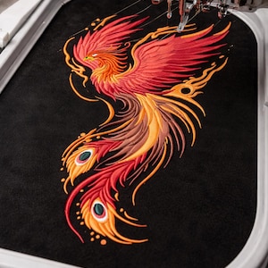 Könnte beinhalten: Eine laufende Stickmaschine, die ein lebendiges Phönix-Design auf schwarzen Stoff stickt. Der Phönix ist mit roten, orangefarbenen und gelben Fäden bestickt und weist komplizierte Federdetails auf. Garnrollen in verschiedenen Farben sind im Hintergrund zu sehen.